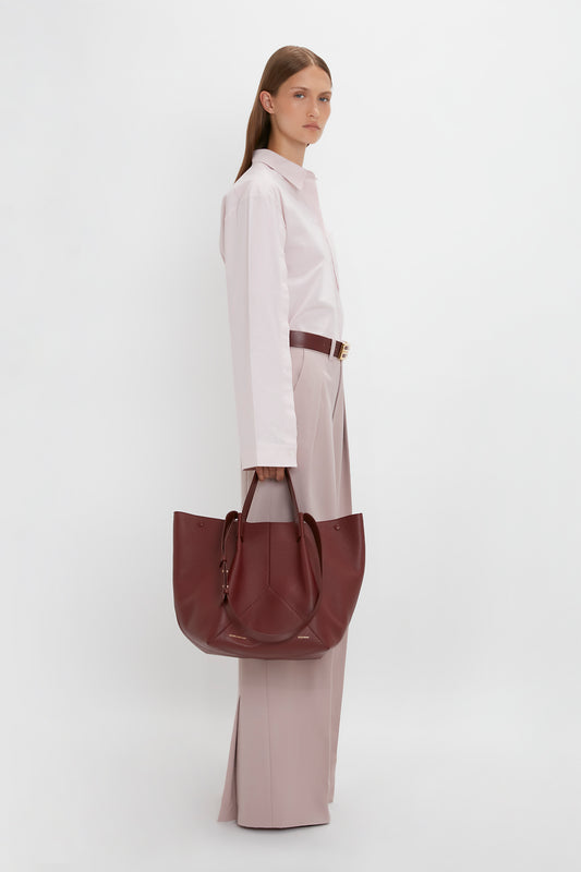 A woman in a beige outfit stands holding a large Victoria Beckham Medium Tote in burgundy leather, viewed from the side against a white background.