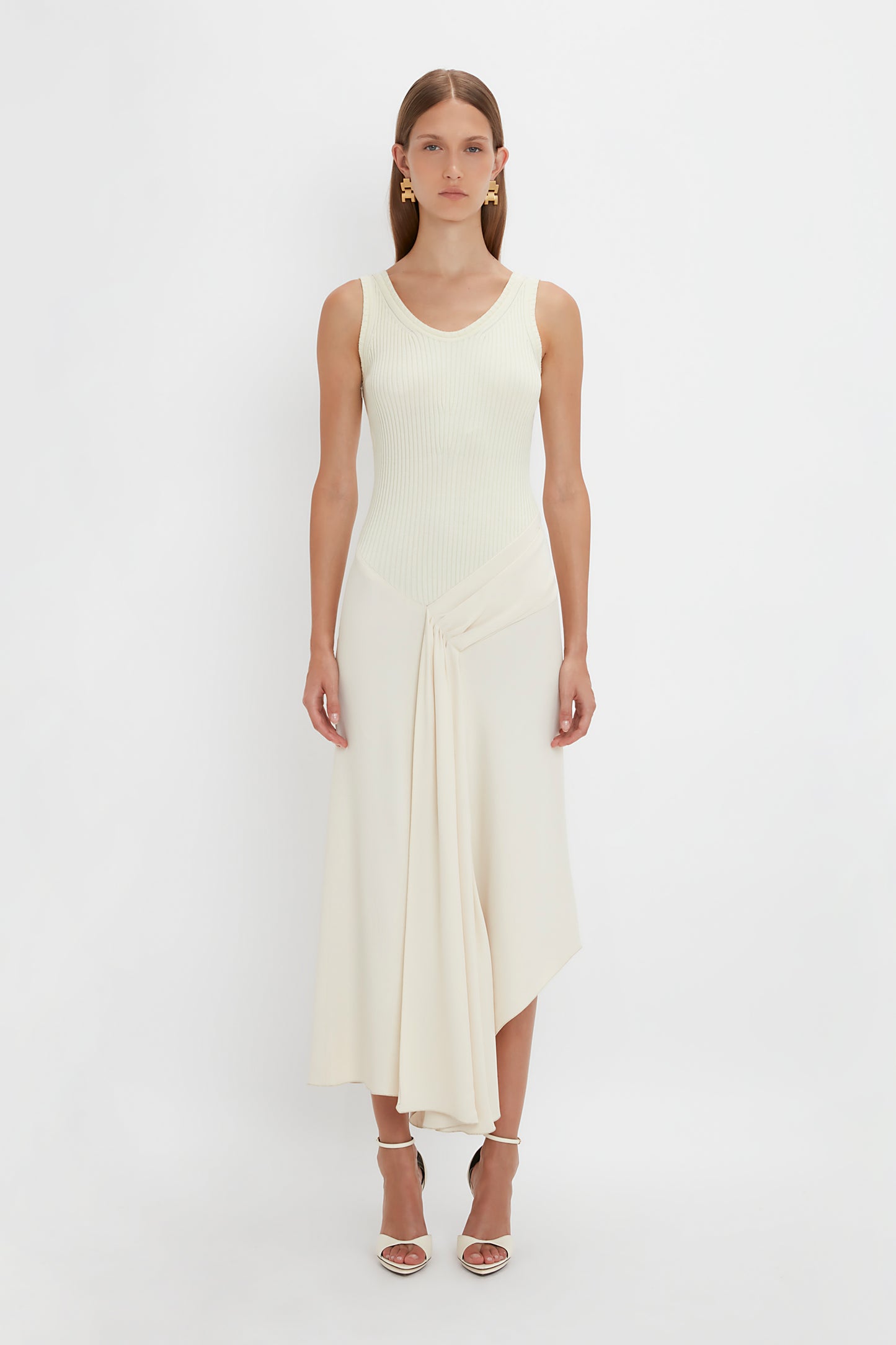 A woman stands against a white background, wearing a Victoria Beckham Sleeveless Tie Detail Dress in Cream and strappy heeled sandals.