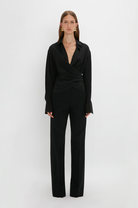 A woman in a Victoria Beckham Wrap Front Blouse In Black and trousers stands against a plain white background.