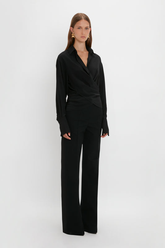 A woman wearing a Victoria Beckham black silk wrap front blouse with wide-leg pants stands against a white background, looking directly at the camera.
