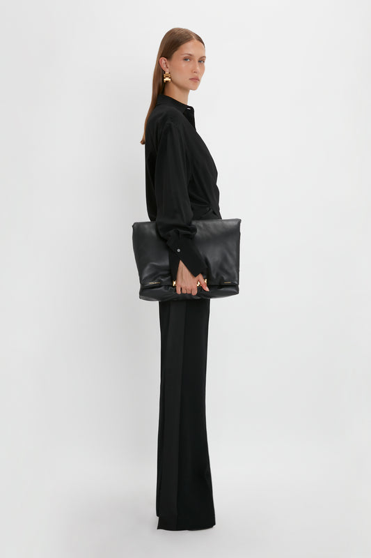 A woman in a Victoria Beckham Wrap Front Blouse In Black holding a large black leather bag, standing sideways against a white background.