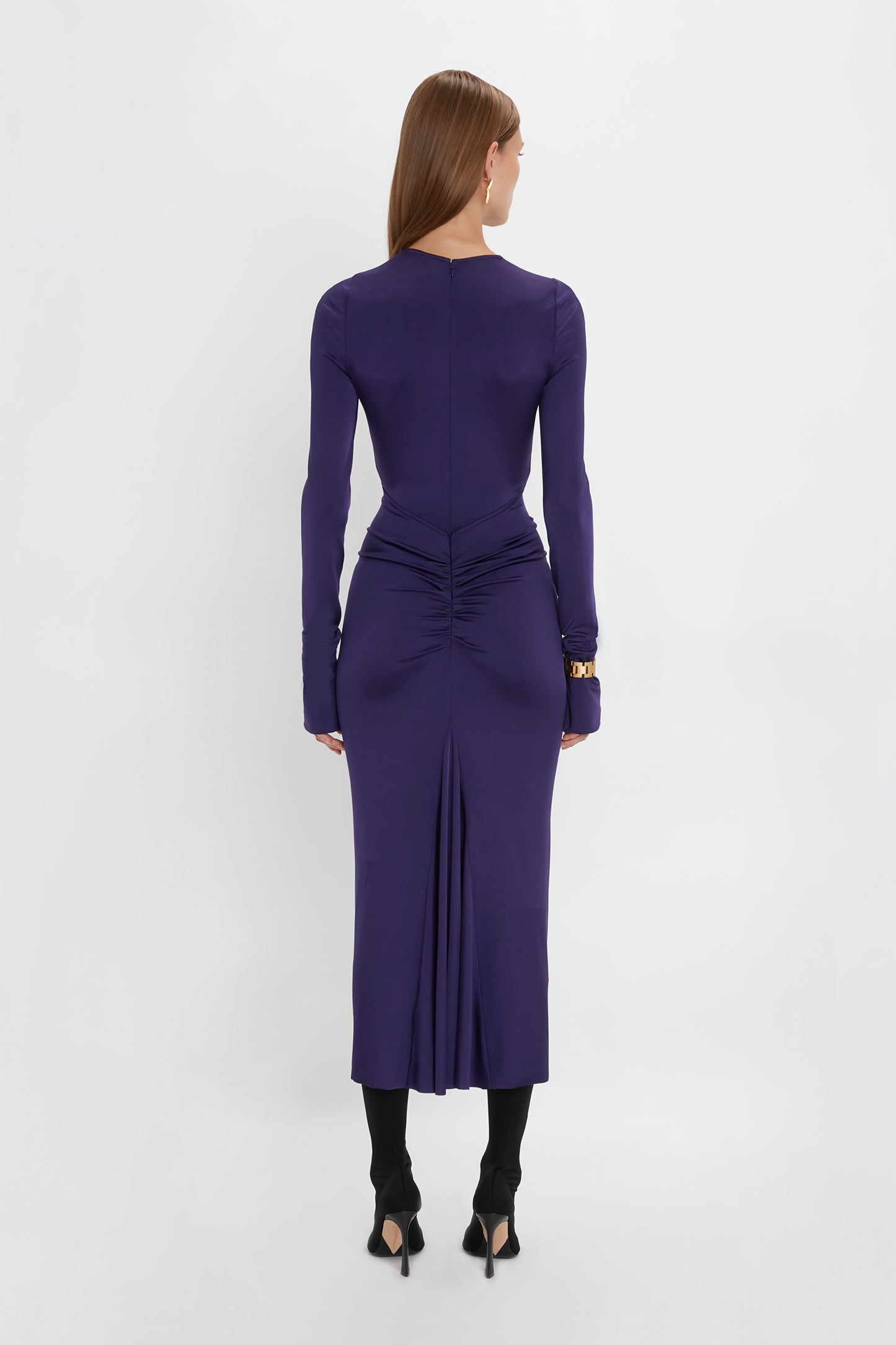 A person stands facing away, wearing a Victoria Beckham Long Sleeve Gathered Midi Dress In Ultraviolet with ruched detailing at the back and a rear slit, showcasing the sensuous and playful design. Paired with black heeled shoes, it's made from body-sculpting stretch fabric for a flawless fit.