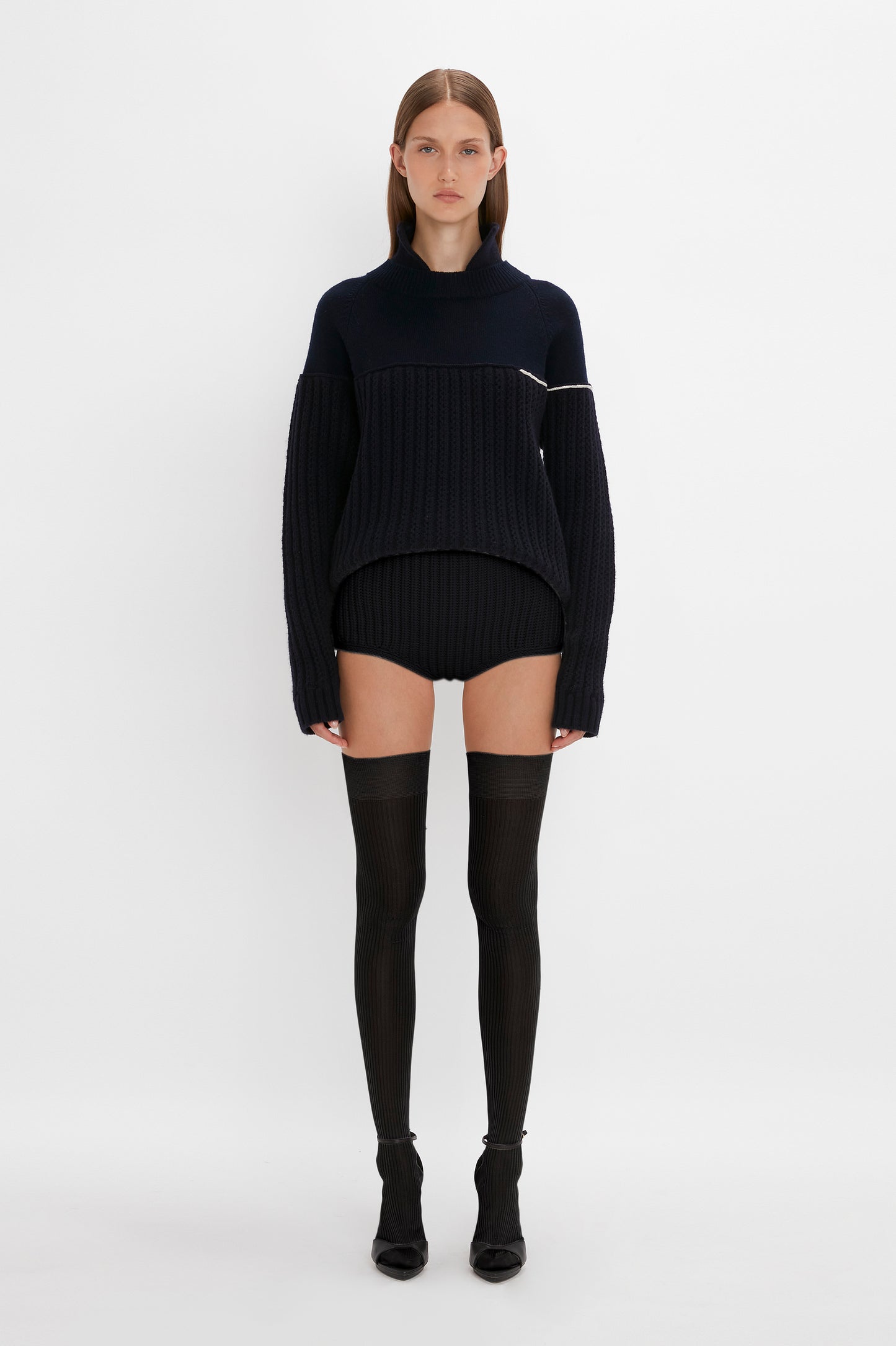 A woman stands center in a studio, wearing a black ribbed sweater dress, Victoria Beckham Exclusive Over The Knee Socks In Black, and short boots, facing forward with a neutral expression.