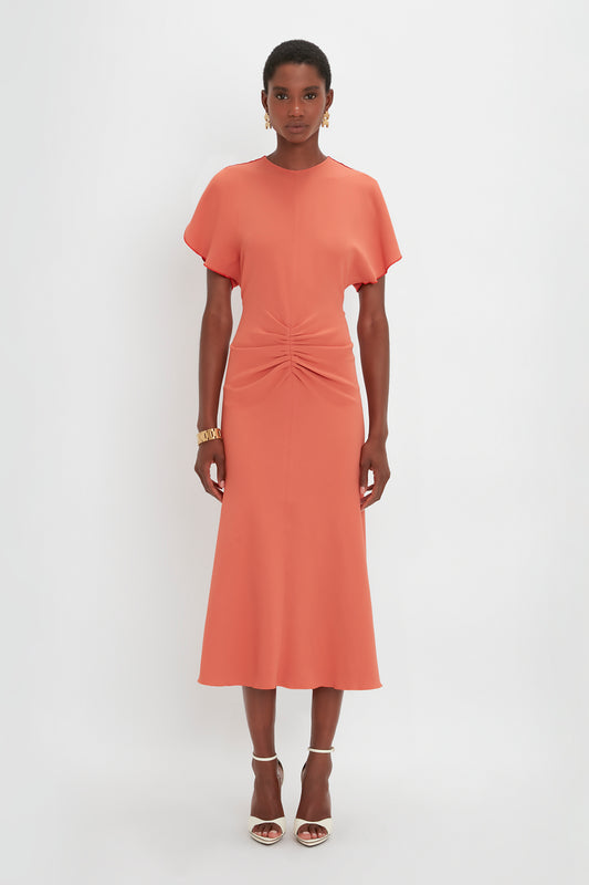 A woman stands against a white background, wearing a Victoria Beckham salmon pink, gathered waist midi dress in papaya, paired with white heels.