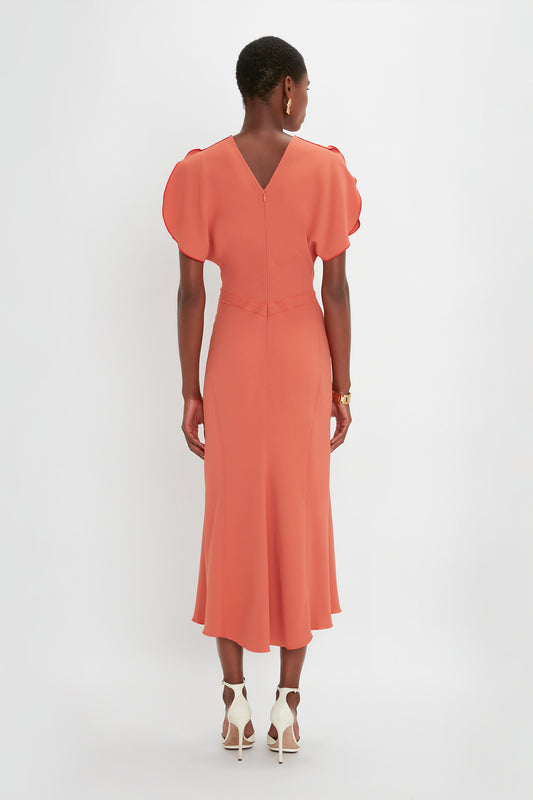 A woman stands with her back to the camera, wearing a Victoria Beckham Gathered Waist Midi Dress In Papaya with puffed sleeves and white high heels.