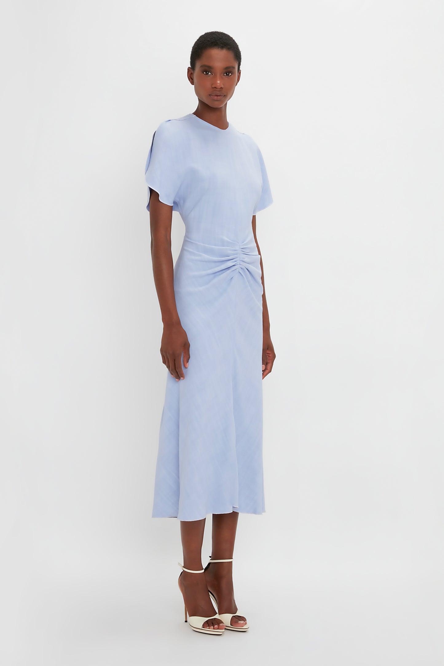 A black woman modeling a Victoria Beckham Gathered Waist Midi Dress In Frost, paired with white pointy toe stiletto sandals, standing against a white background.