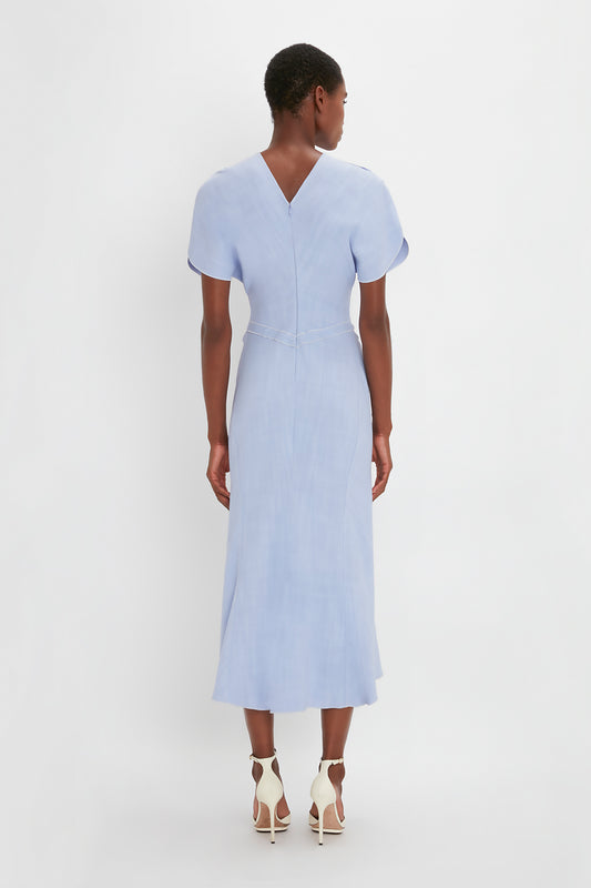 Woman from behind, wearing a Victoria Beckham light blue Gathered Waist Midi Dress In Frost and white pointy toe stiletto sandals, standing against a plain white background.