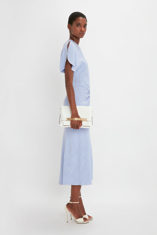 A woman in a Victoria Beckham light blue Gathered Waist Midi Dress In Frost and white pointy toe stiletto sandals, holding a white handbag, poses sideways against a white background.