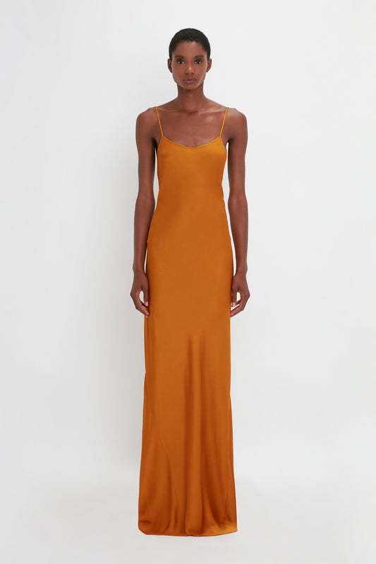 A black woman stands against a white background, wearing a long, camisole-slip mustard-yellow dress with thin straps by Victoria Beckham.