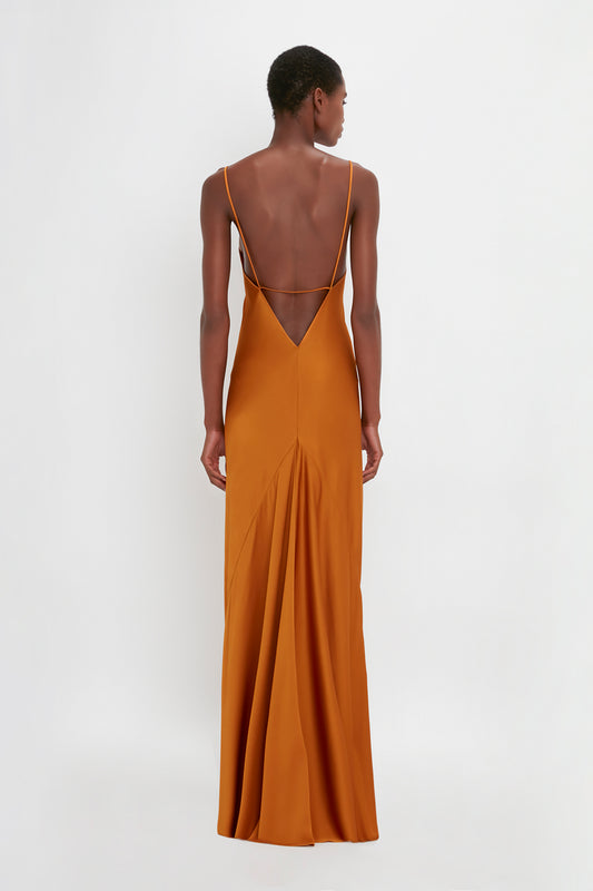 A woman standing with her back to the camera, wearing an elegant, deep orange Victoria Beckham floor-length cami dress in ginger with crisscross straps.