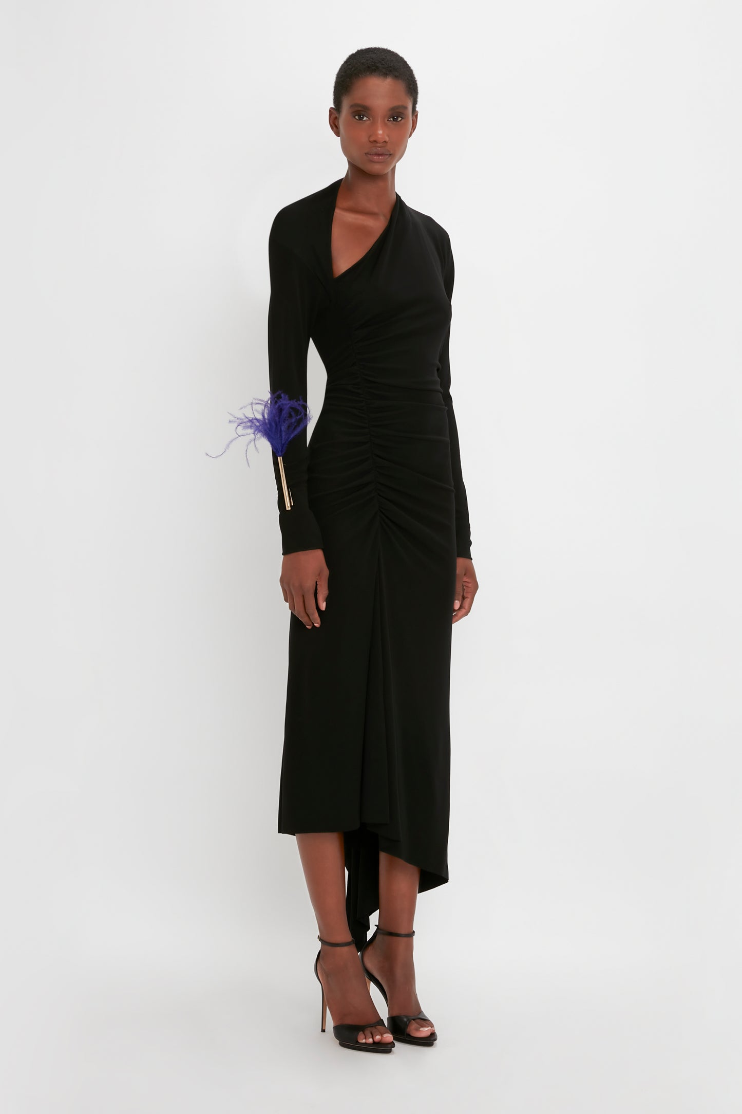 A woman models a Victoria Beckham black gown with ruched details and a blue feather accent on the wrist, paired with black strappy heels.