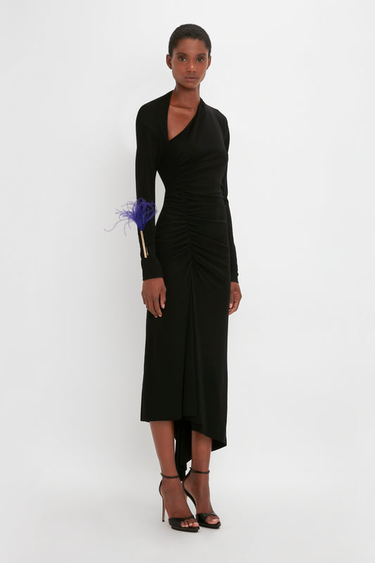 A woman models a Victoria Beckham black gown with ruched details and a blue feather accent on the wrist, paired with black strappy heels.