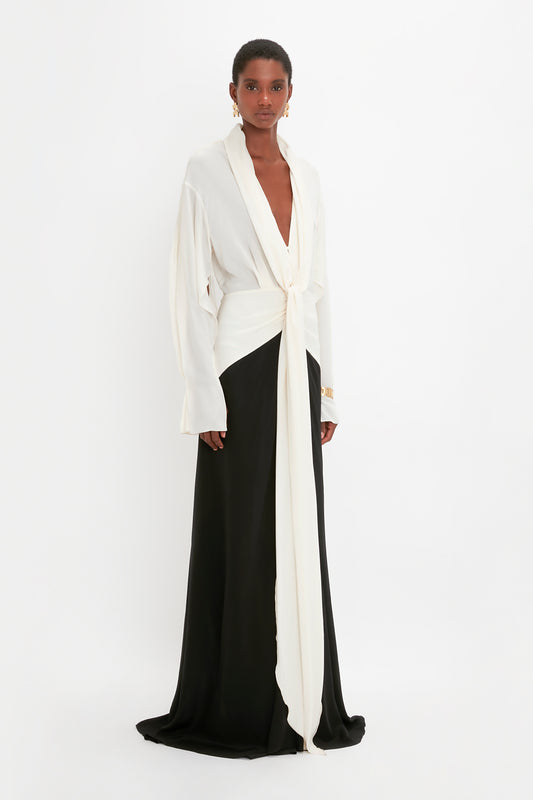 A woman stands wearing a Victoria Beckham Tie Detail Gown in Vanilla-Black featuring a long black evening gown and a white wrapped top with long sleeves and a plunging neckline.