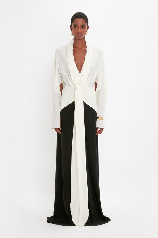 A woman stands facing the camera, wearing an elegant Tie Detail Gown In Vanilla-Black by Victoria Beckham with a deep v-neckline and long sleeves. She has a gold bracelet on her left wrist.