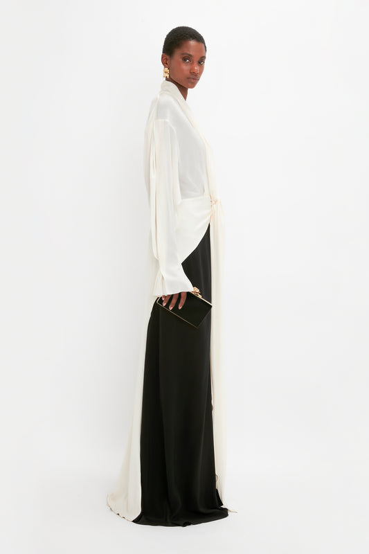 A woman in a Victoria Beckham Tie Detail Gown In Vanilla-Black with a deep-V neckline clutching a small black purse, standing against a white background.