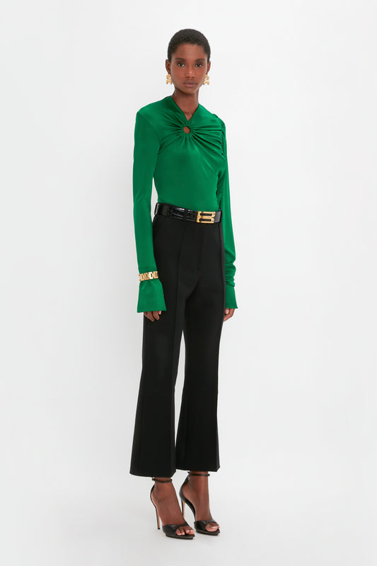 A woman in a vibrant green blouse and black trousers, accessorized with an Exclusive Jumbo Chain Bracelet in Brushed Gold from Victoria Beckham UK, stands facing the camera against a white background.