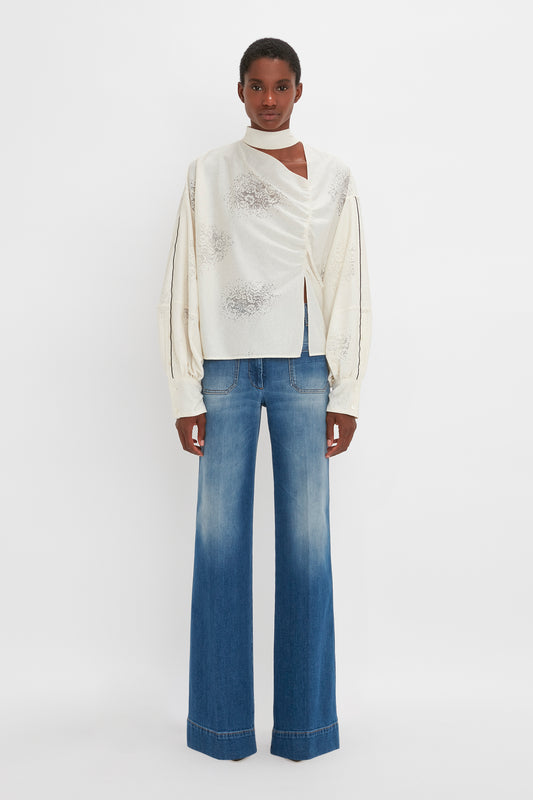 A woman standing against a white background, wearing a unique Asymmetric Gather Detail Top in Cream by Victoria Beckham with blouson sleeves and blue flared jeans.