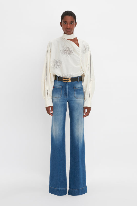 A woman in a cream, 100% silk Asymmetric Gather Detail Top by Victoria Beckham with blouson sleeves and blue wide-leg jeans standing against a white background.