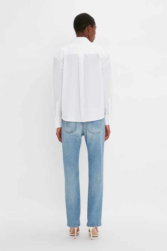 Rear view of a woman wearing a Victoria Beckham Cropped Long Sleeve Shirt in White and blue jeans, standing against a white background.