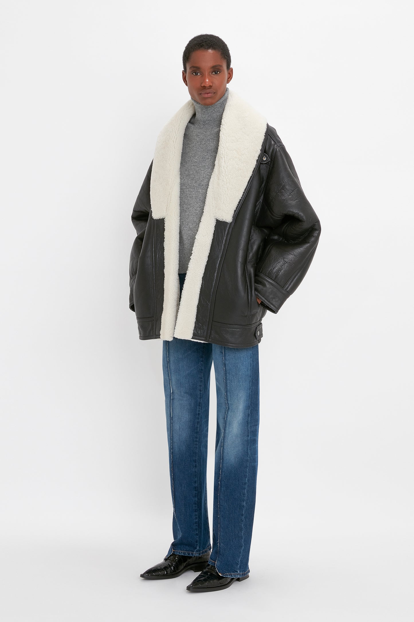 Person standing against a white background wearing a cream and black polo neck jumper, the Victoria Beckham Shearling Coat In Monochrome, and blue jeans.