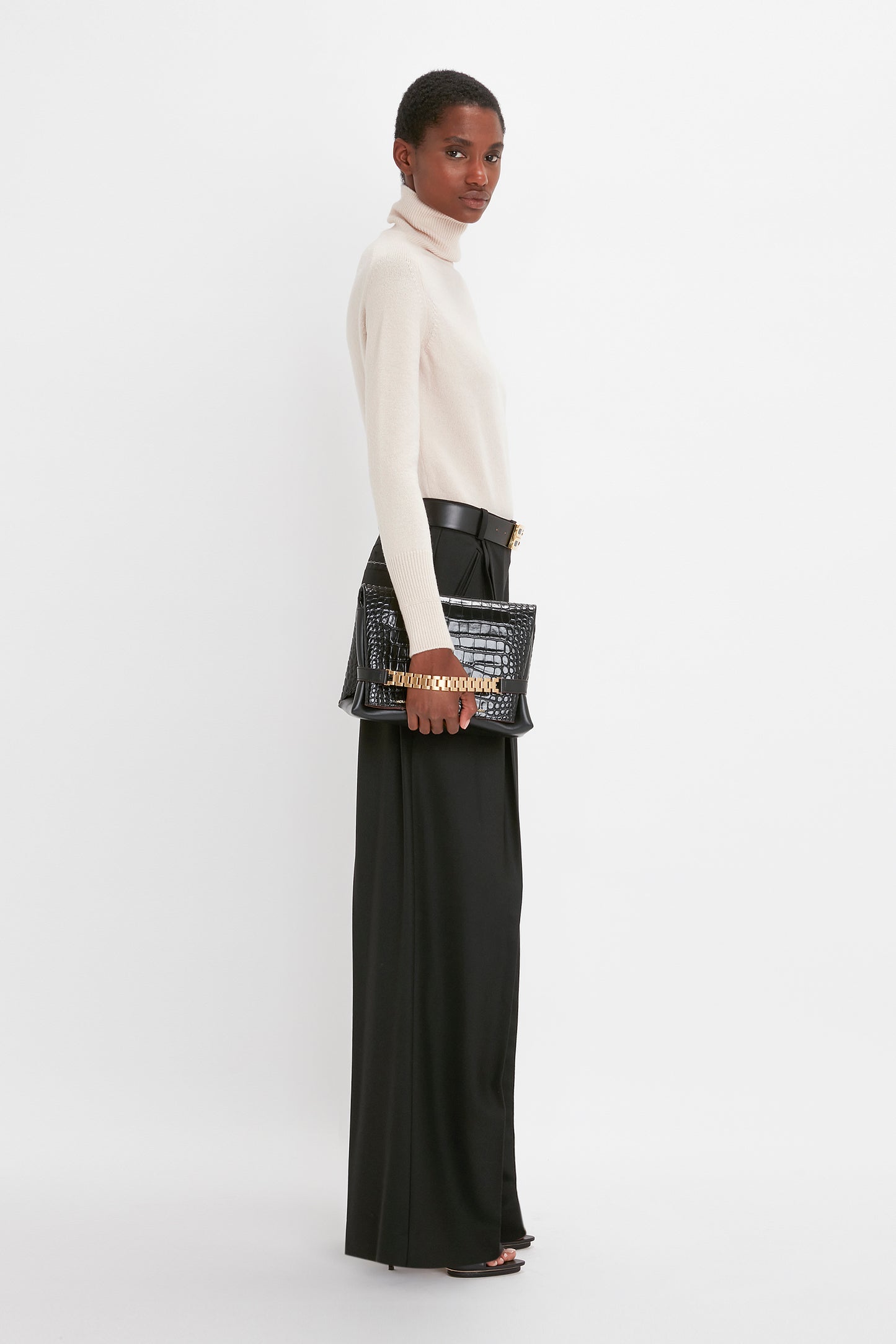 A woman in a cream Victoria Beckham polo neck jumper and black wide leg trousers, holding a black studded handbag, looking over her shoulder against a white background.