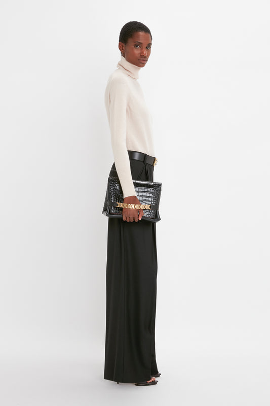 A woman in a cream Victoria Beckham polo neck jumper and black wide leg trousers, holding a black studded handbag, looking over her shoulder against a white background.