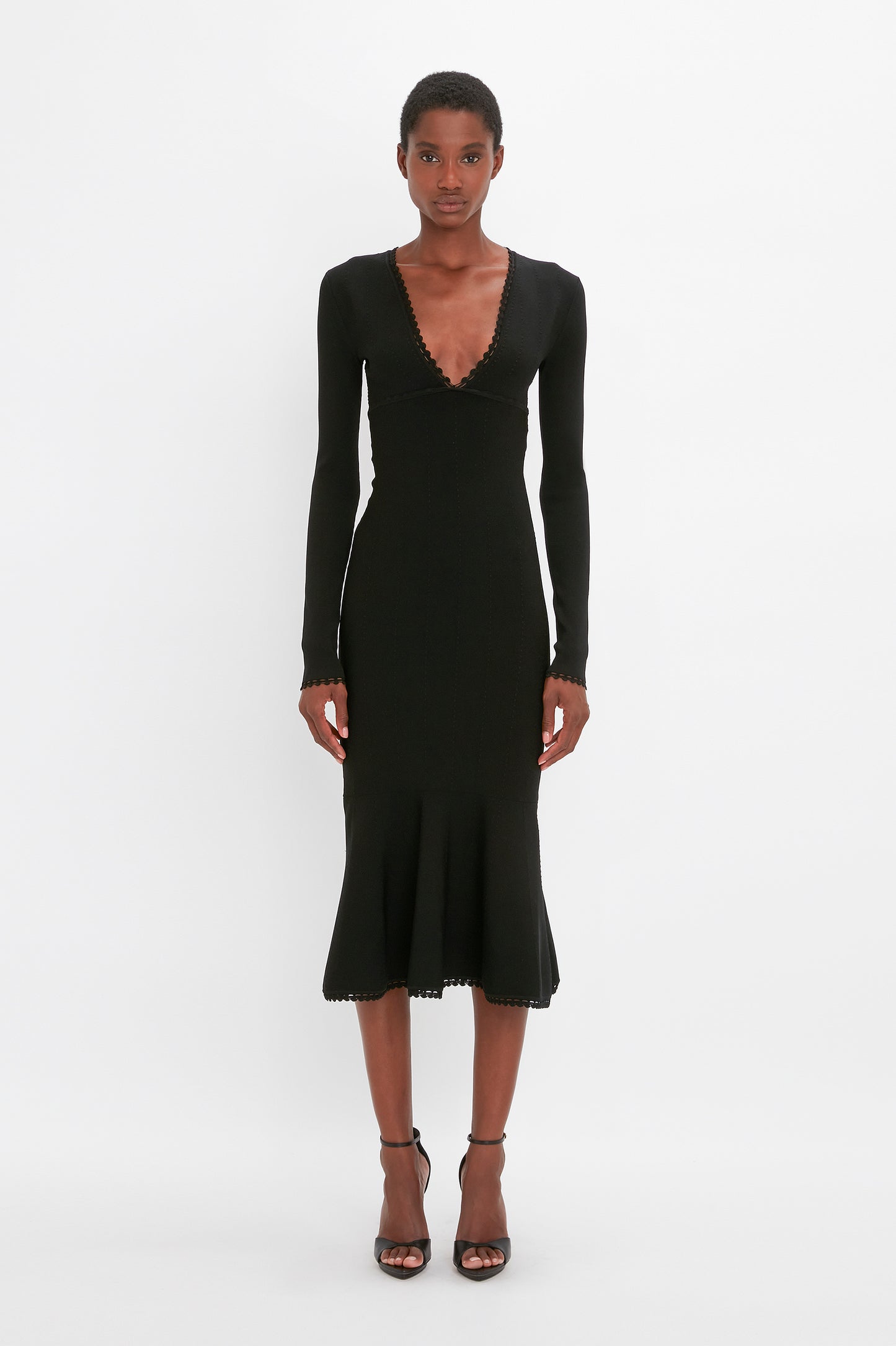 A person stands against a plain white background, wearing the VB Body Long Sleeve V Neck Dress In Black by Victoria Beckham. They are also wearing black strappy heels.