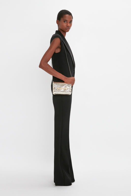 A woman in a sleek black sleeveless jumpsuit stands sideways, holding a Victoria Beckham Mini Chain Pouch With Long Strap In Gold Leather, looking over her shoulder.