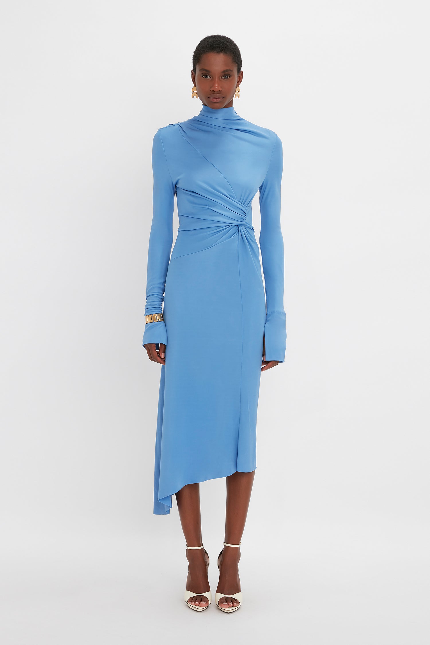 A woman in a Victoria Beckham High Neck Asymmetric Draped Dress In Oxford Blue with a twist detail at the waist, standing against a white background.