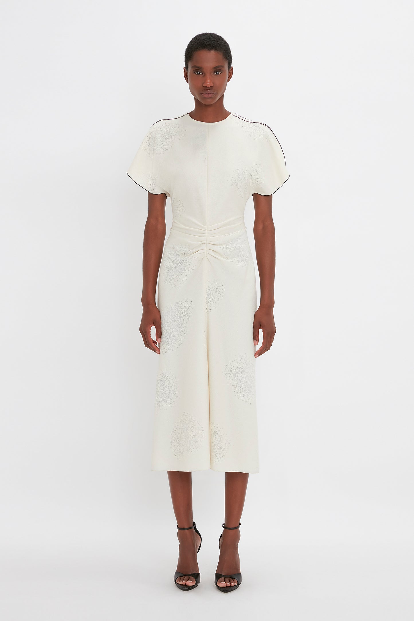 A woman stands against a white background, modeling a cream Victoria Beckham gathered waist midi dress with short sleeves and delicate embroidery, paired with pointy toe stiletto sandals.