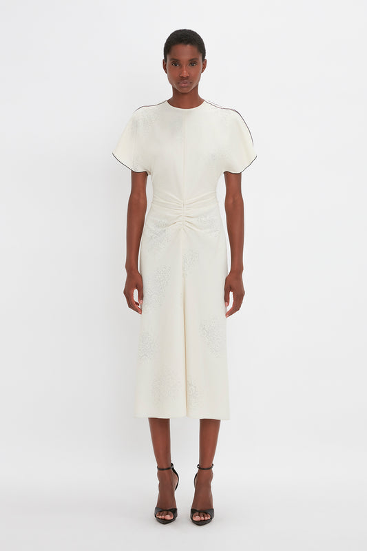 A woman stands against a white background, modeling a cream Victoria Beckham gathered waist midi dress with short sleeves and delicate embroidery, paired with pointy toe stiletto sandals.
