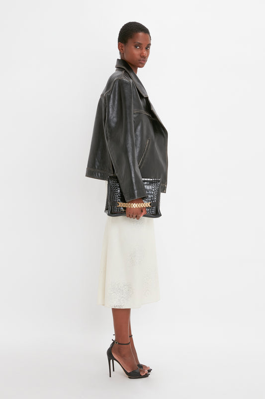 A woman in a stylish oversized leather jacket and elegant white skirt posing sideways on a plain background.