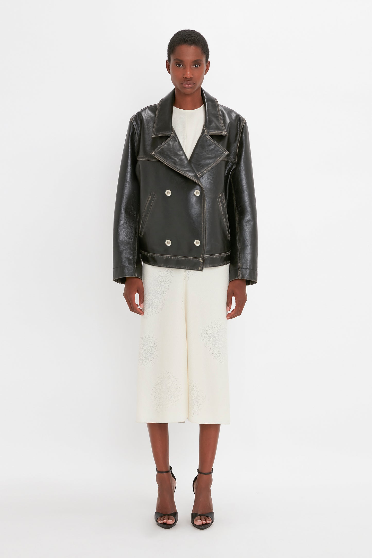 A person standing against a plain background, wearing a white outfit with an Oversized Leather Jacket In Black by Victoria Beckham and black sandals.