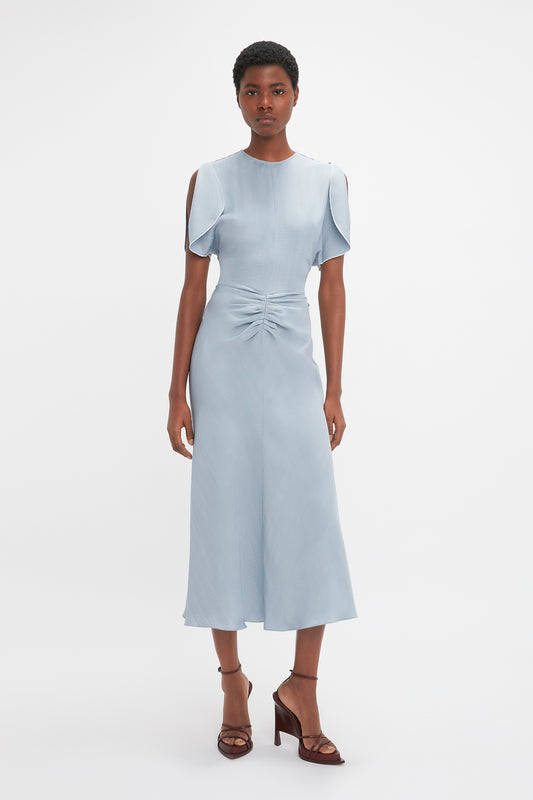 A woman stands on a plain background wearing a light blue Exclusive Gathered Waist Midi Dress In Pebble by Victoria Beckham with shoulder cut-outs and a gathered detail at the waist, paired with dark heels.
