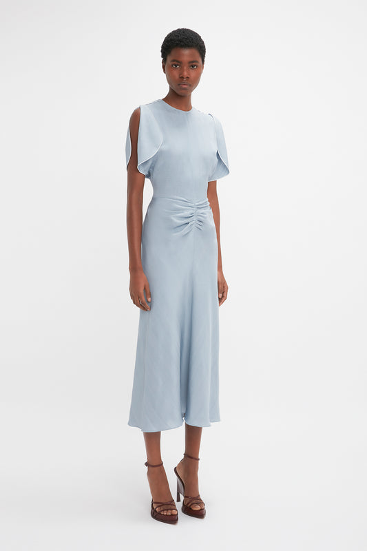 A woman in a light blue Victoria Beckham Exclusive Gathered Waist Midi Dress In Pebble with ruffled sleeves, standing against a plain white background.