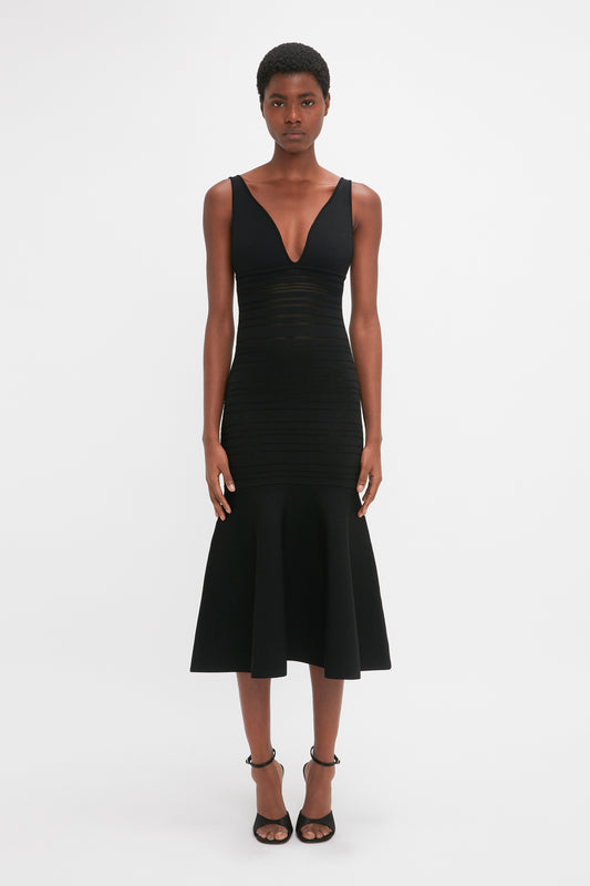 A black woman standing against a white background, wearing a Victoria Beckham black sleeveless fit-and-flare dress with a v-neckline and midi-length skirt, paired with black heels.