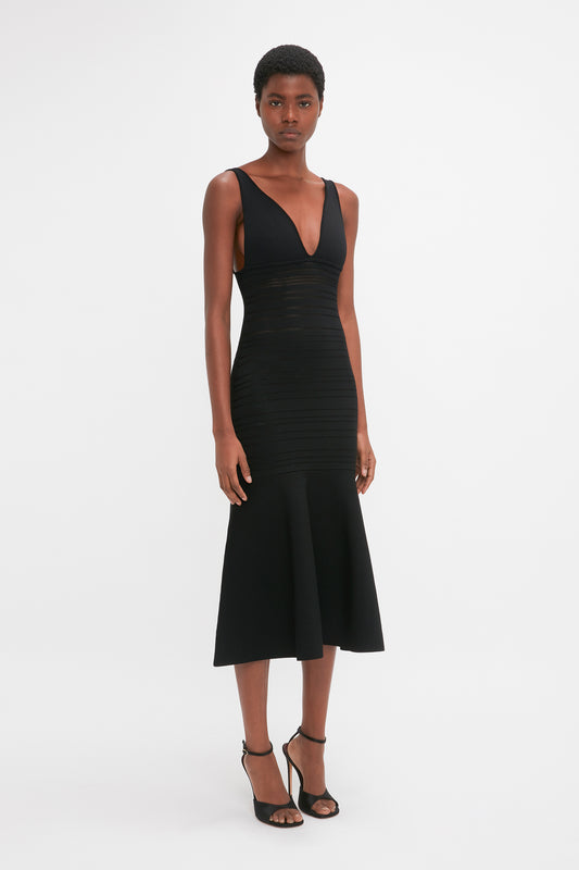 A black woman wearing a Victoria Beckham Frame Detail Sleeveless Dress in black, with a plunging neckline and midi-length skirt, standing against a white background.