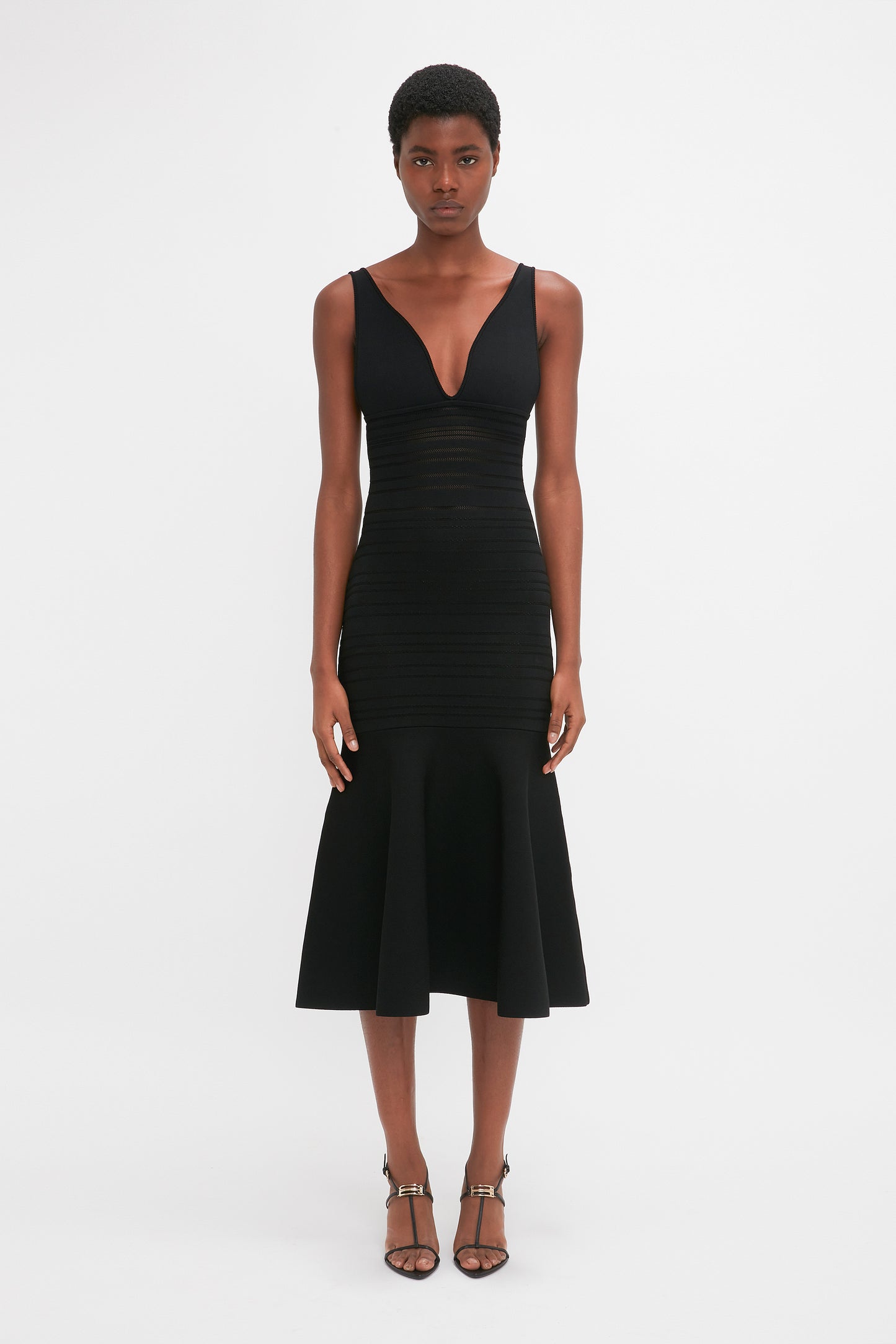 A person stands facing forward wearing a sleeveless, knee-length black dress with a deep V-neck. They are also wearing black strappy heels featuring a distinctive V-shaped sculptural heel: the Frame Detail Sandal In Black Leather by Victoria Beckham. The background is plain white.