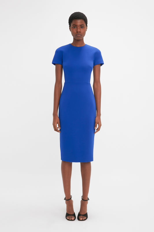 A woman in a Victoria Beckham Palace Blue fitted t-shirt dress and black heels stands against a white background, looking directly at the camera.