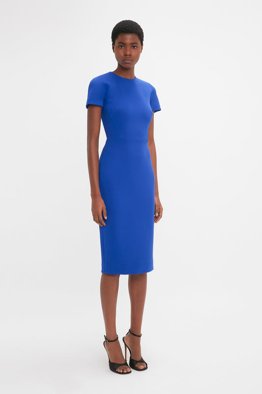 African American woman standing in a studio, wearing a Victoria Beckham fitted T-shirt dress in palace blue wool crepe knee-length dress and black high-heeled sandals.