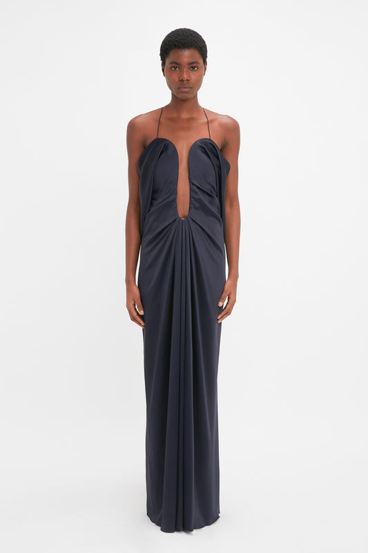A black woman stands facing the camera, wearing a Victoria Beckham navy blue sleeveless evening gown with a cut-out design at the bust.