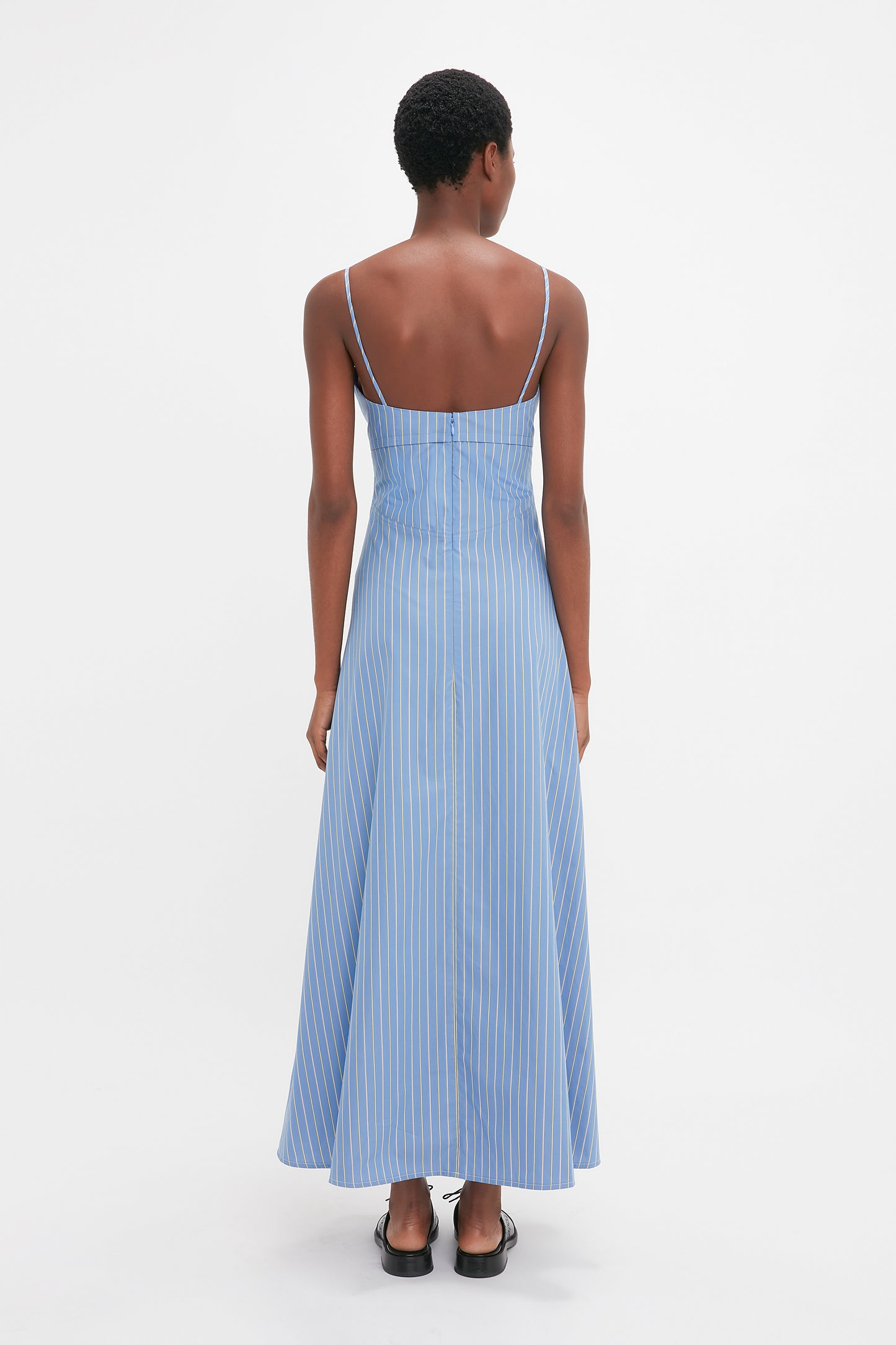 A person with short hair is standing and facing away, wearing a blue and white striped cotton **Cami Fit And Flare Midi In Steel Blue** with spaghetti straps, paired with black flat lace-up mules by **Victoria Beckham**. The background is plain white.