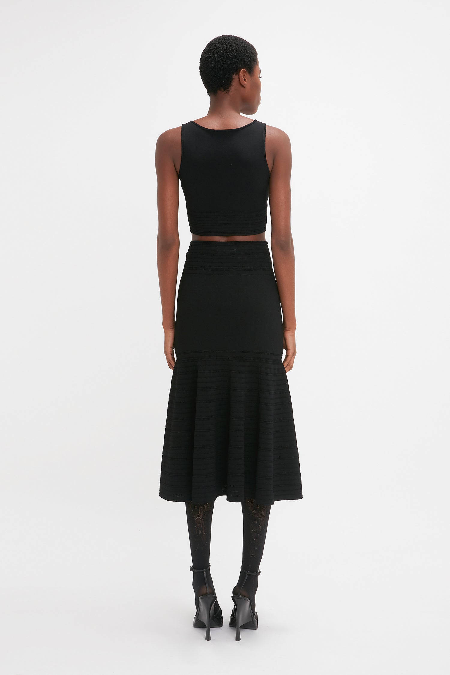 A woman from behind, in a black Victoria Beckham Frame Detail Sleeveless Top and pleated skirt with a cropped hemline, paired with high heels and patterned tights, standing against a white background.