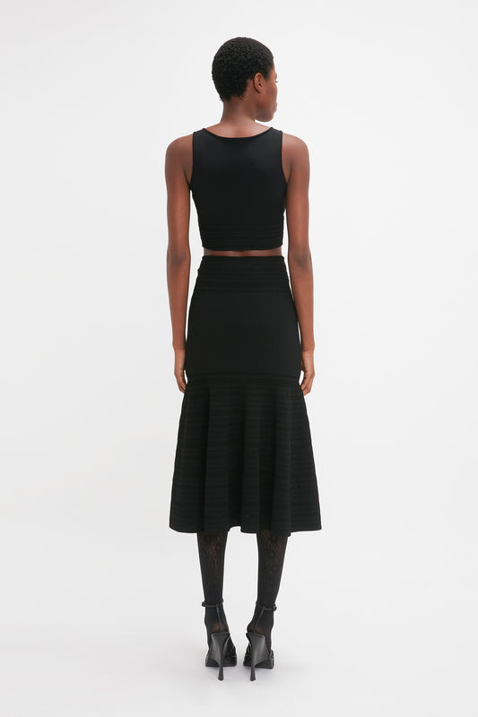 A woman from behind, in a black Victoria Beckham Frame Detail Sleeveless Top and pleated skirt with a cropped hemline, paired with high heels and patterned tights, standing against a white background.