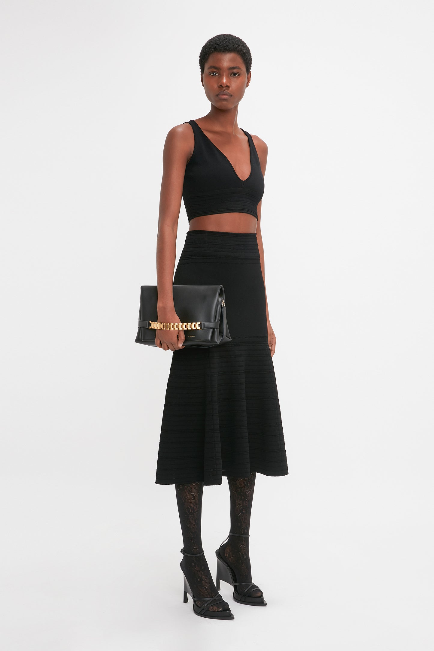 A person is standing against a plain white background, wearing a black sleeveless top, the Victoria Beckham Fit And Flare Midi Skirt In Black, black high-heeled shoes, and holding a black handbag with contrasting stitching.