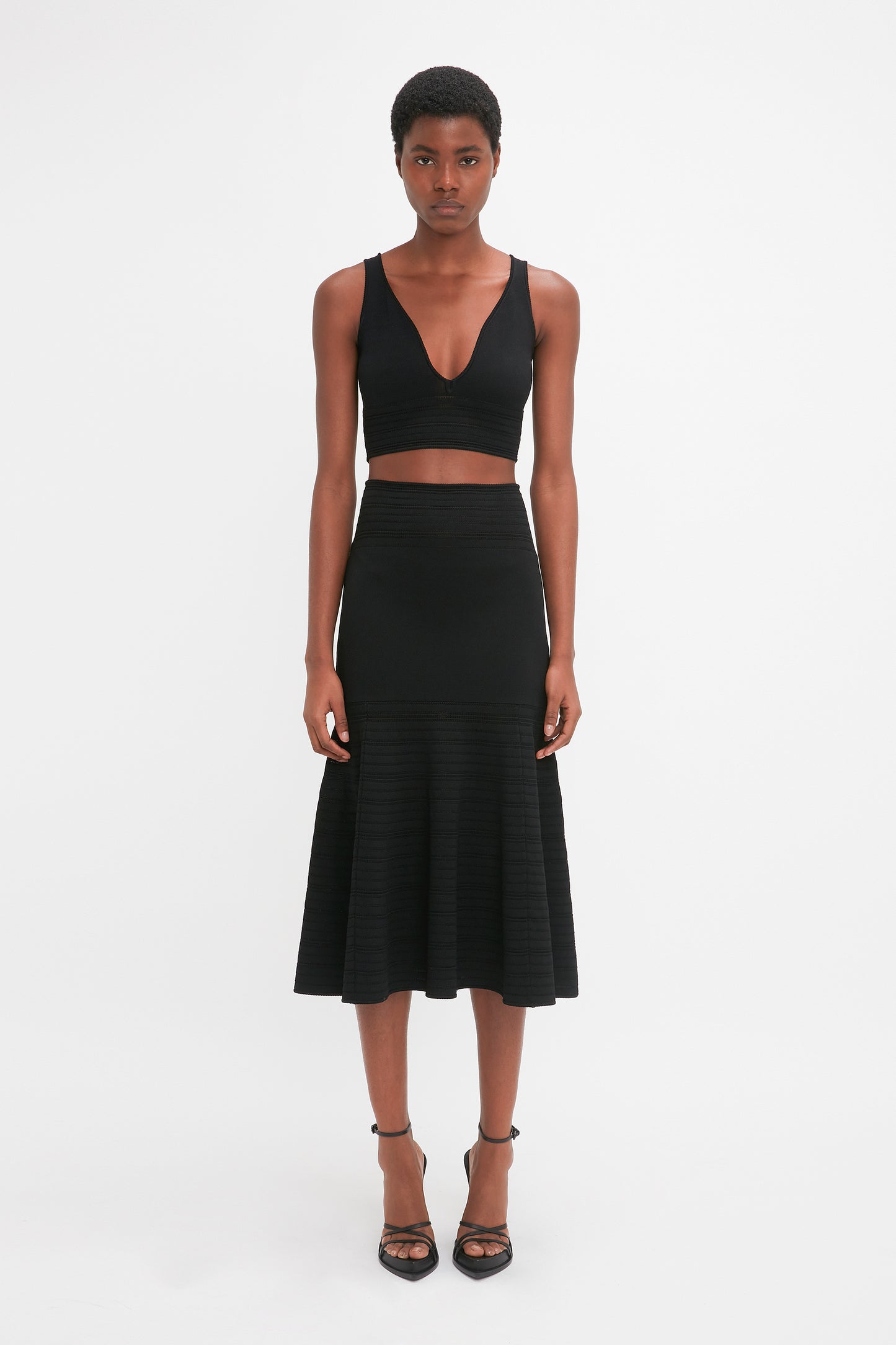 A person stands against a white background, wearing a black sleeveless top and the Fit And Flare Midi Skirt In Black by Victoria Beckham made from mid-weight stretch knit fabric, paired with black heels. They are looking directly at the camera.