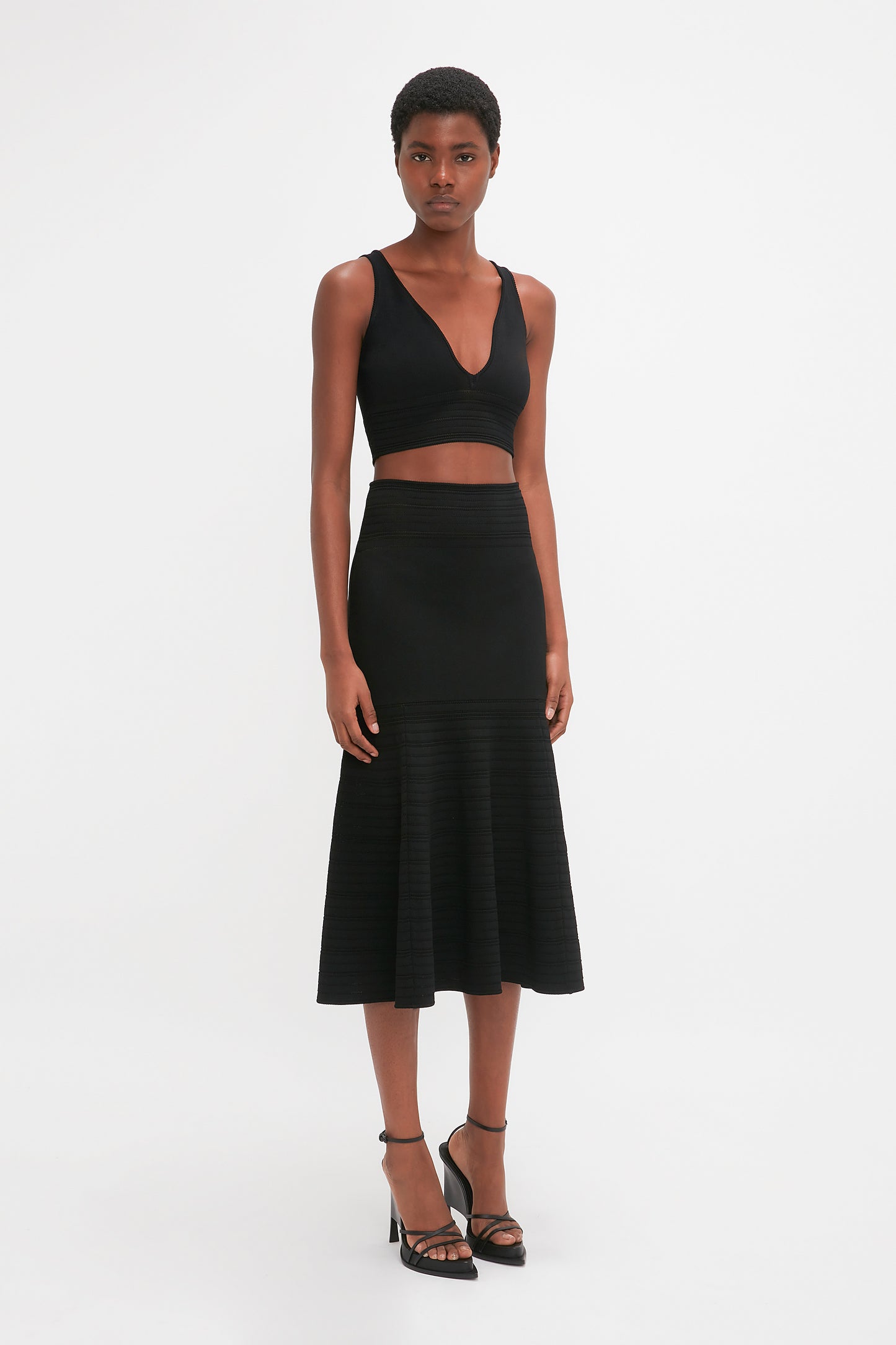A person with short hair stands wearing a black sleeveless crop top and a waist-defining Victoria Beckham Fit And Flare Midi Skirt In Black made of mid-weight stretch knit, paired with black high-heeled sandals, against a plain white background.