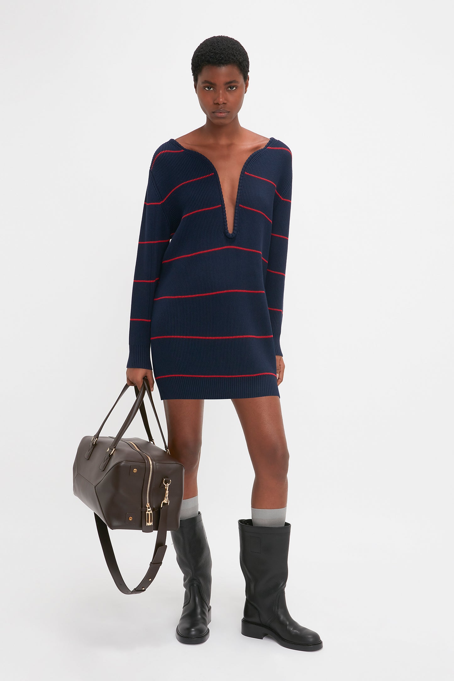A person in a Frame Detail Jumper Dress In Navy-Red by Victoria Beckham, black boots, and gray socks stands holding a brown bag, embodying the SS24 runway style with its chic mid-weight knit design.