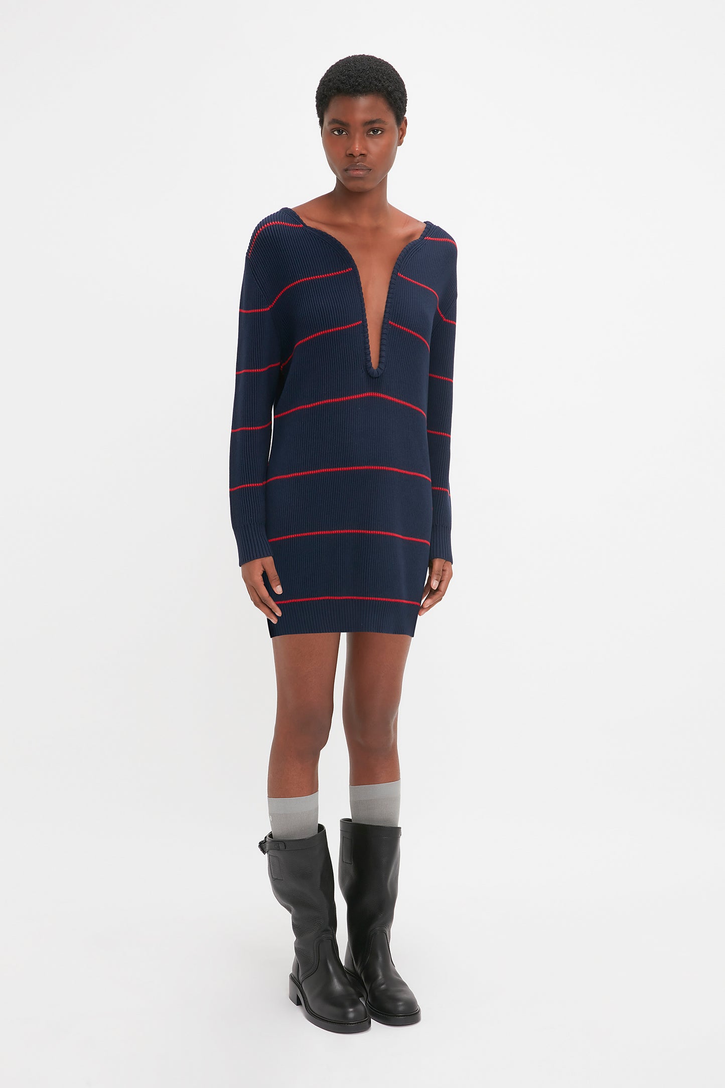 A person in a long-sleeved, Frame Detail Jumper Dress in Navy-Red by Victoria Beckham with a deep V-neckline, gray socks, and black boots, standing against a white background. The mid-weight knit fabric adds texture and style reminiscent of the SS24 runway elegance.