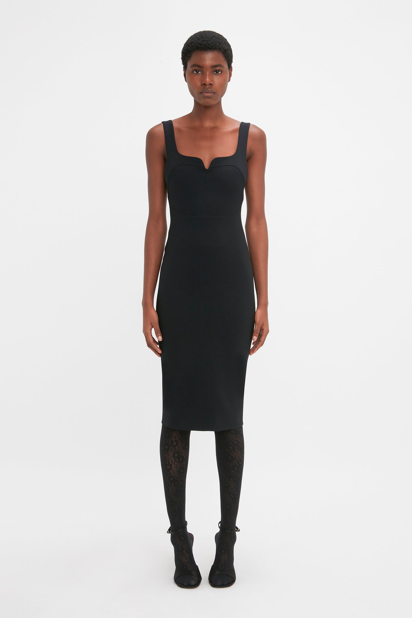 Person standing against a plain white background, wearing a Sleeveless Fitted T-Shirt Dress In Black by Victoria Beckham, patterned black tights, and black shoes. The expression is neutral.
